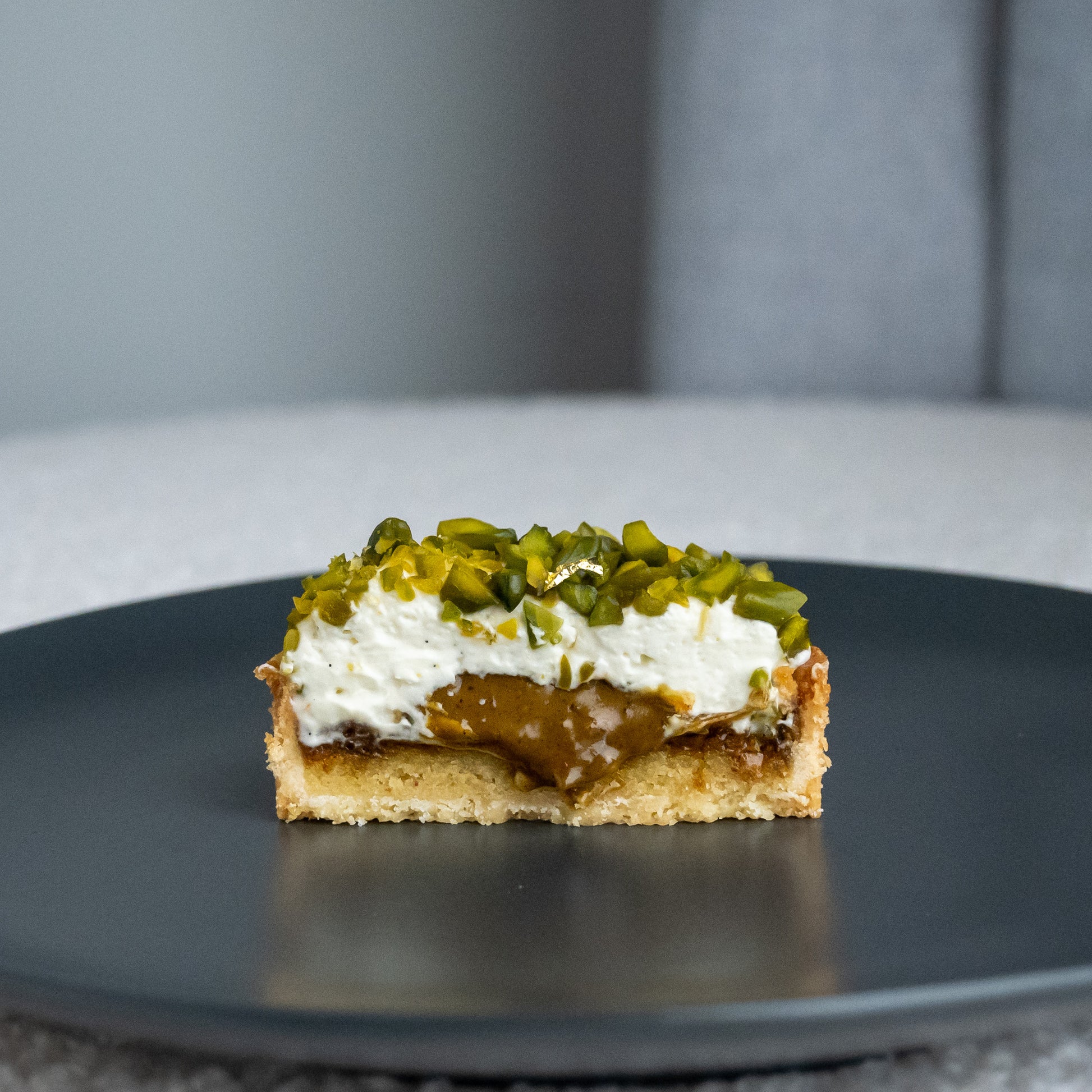 A cross-section view of our best-selling Pistachio Tart, revealing layers of crispy tart base, frangipane cream, crunchy pistachio praliné, and a liquid center of praliné. The tart is topped with a dollop of vanilla chantilly cream and sprinkled with chopped Iranian pistachios. The oozing liquid center adds an extra touch of decadence to this already indulgent treat.