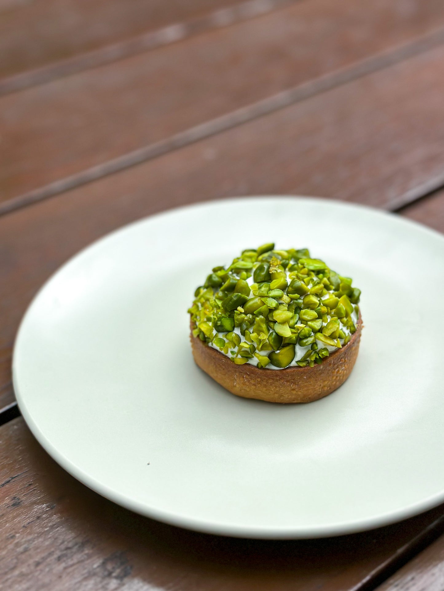 Our popular Pistachio Tart - with a crispy tart base, frangipane cream, crunchy pistachio praliné layer, liquid center of praliné, vanilla chantilly cream, and topped with chopped Iranian pistachios. The tart is rich in color, with layers of green, yellow, and beige, and is cut to show the different layers of the filling.
