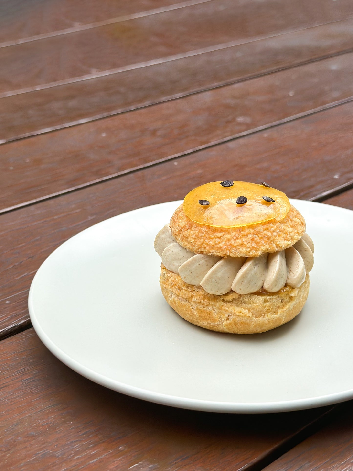 A close-up of a golden-brown choux pastry puff. The choux puff is sliced horizontally, revealing a filling of smooth banana cream and tangy passion fruit confit inside a mini choux. The top of the choux puff is garnished with a spoonful of sweet banana compote.